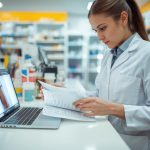 Farmacêutico de jaleco confere receituário impresso enquanto utiliza notebook para registrar no Sistema Nacional de Controle de Receituários (SNCR).